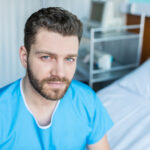 A man with short brown hair and a beard, wearing blue scrubs, sits beside a hospital bed in a medical room, possibly discussing colorectal cancer treatment. Medical equipment is visible in the background as he looks at the camera with a neutral expression.