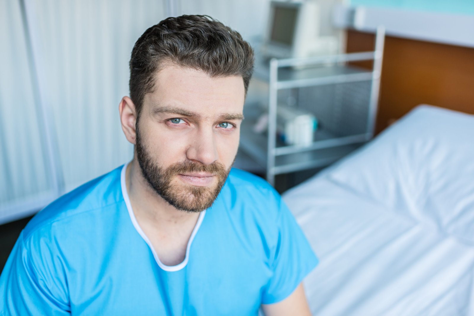 A man with short brown hair and a beard, wearing blue scrubs, sits beside a hospital bed in a medical room, possibly discussing colorectal cancer treatment. Medical equipment is visible in the background as he looks at the camera with a neutral expression.