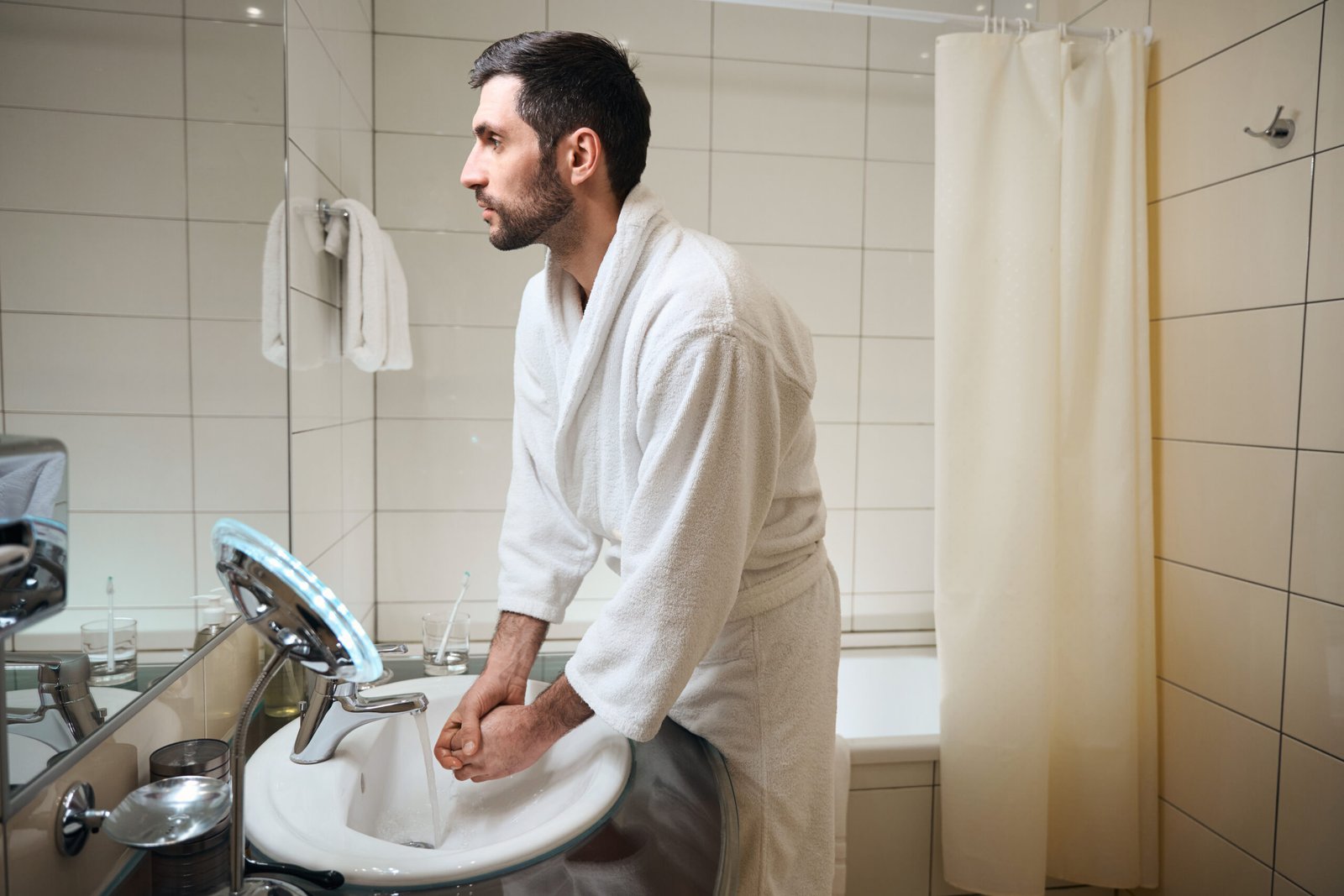 A man in a white dressing gown stands at a bathroom sink, washing his hands. The bathroom has white tiled walls, a mirror, a towel, and a shower curtain in the background.
