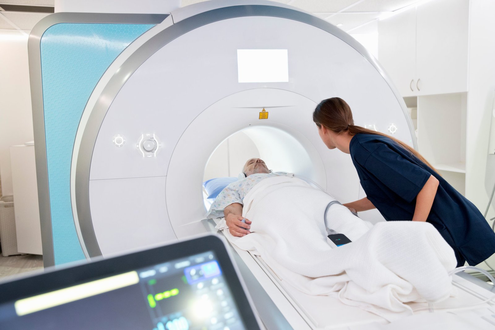A medical professional assists an elderly patient lying on a bed as they are positioned for an MRI scan to check for prostate cancer, with medical equipment visible in the foreground.