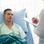A man in a hospital bed wearing a light green gown looks at a testicular cancer doctor, who is standing and writing on a clipboard. The setting is bright with natural light coming through a window.