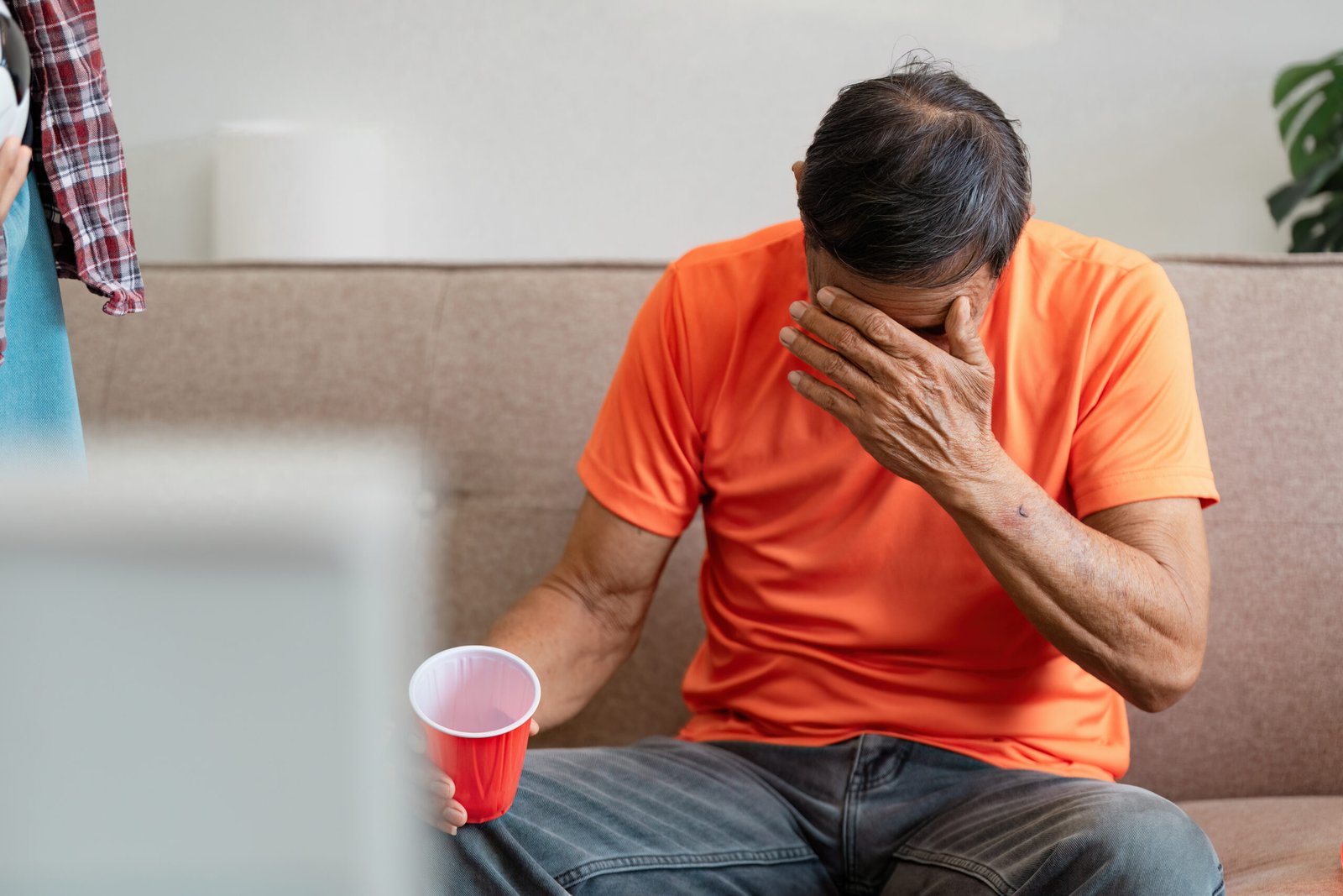 A man in an orange shirt sits on a sofa, holding a red cup in one hand and covering his face with the other, appearing distressed—possibly after receiving news about prostate cancer. Another person stands nearby, partially visible.