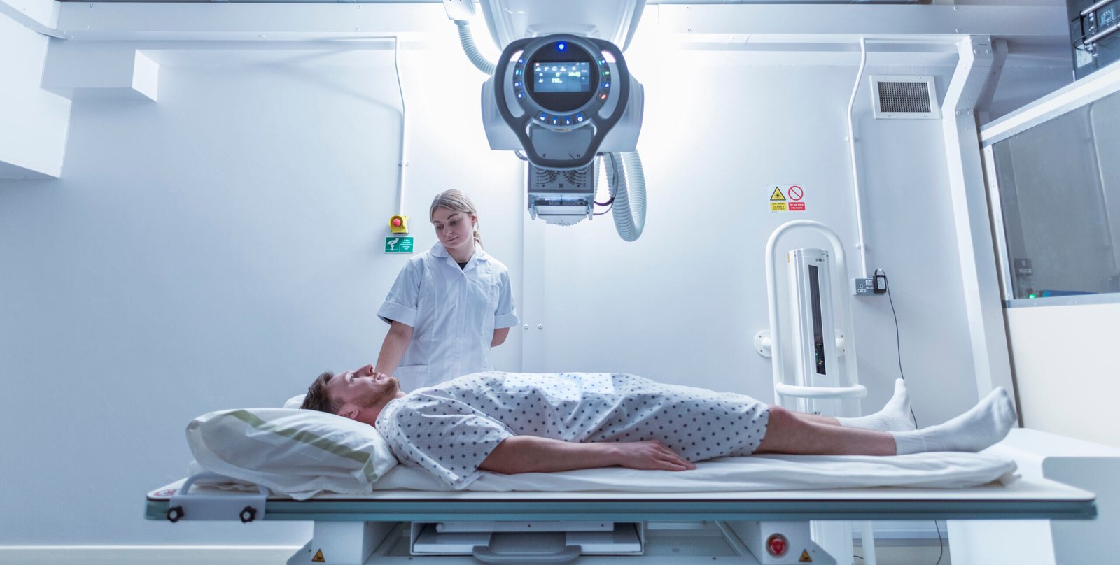 A patient in a hospital gown lies on a bed under a large medical imaging machine, whilst a healthcare worker in a white uniform stands nearby in a bright, modern hospital ward.