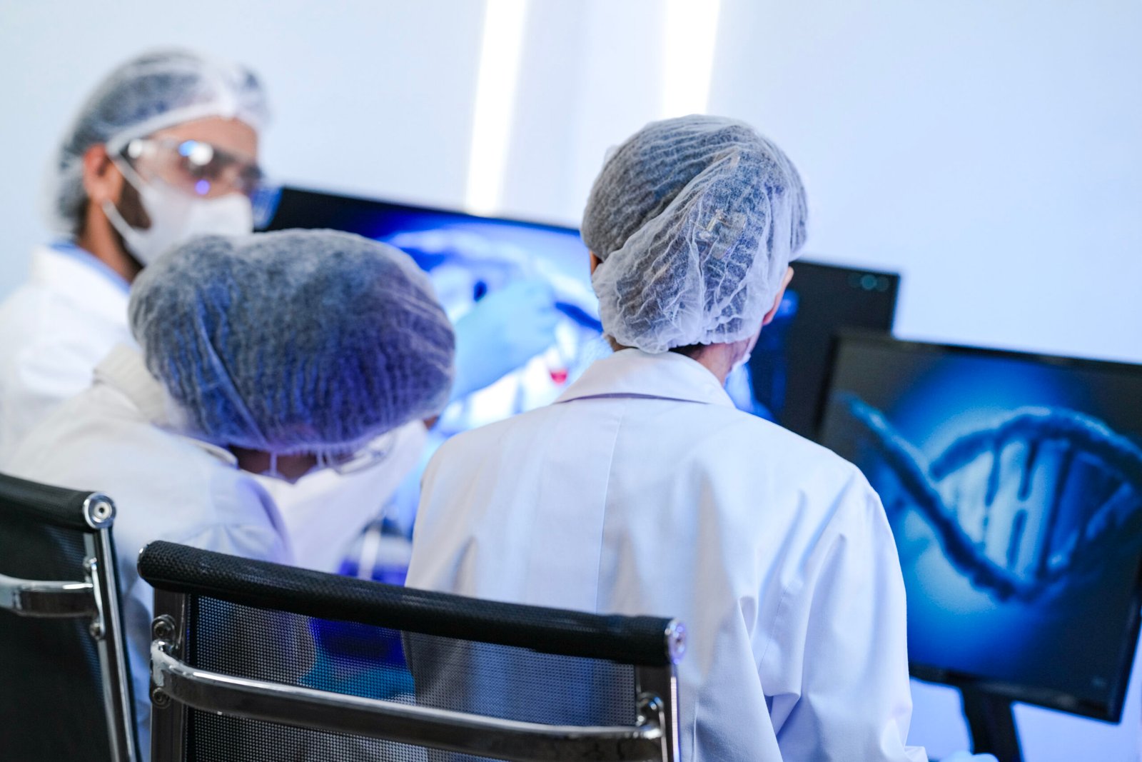 Three scientists wearing lab coats, hairnets, and masks are seated at computer monitors displaying DNA graphics, collaborating on research into treatment options for prostate cancer in a modern laboratory setting.