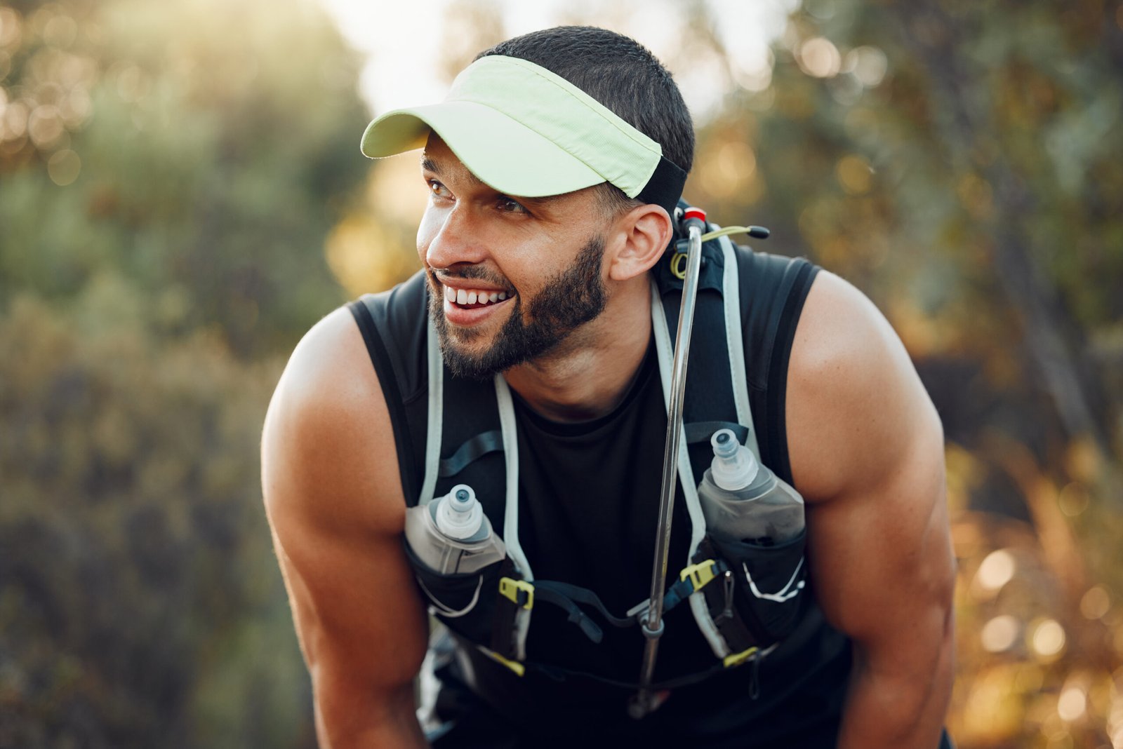 A smiling man with a beard wears a sun visor and hydration vest with water bottles, leaning forward outdoors on a sunny day after running or hiking—staying active while exploring treatment options for prostate cancer. Trees and sunlight fill the background.