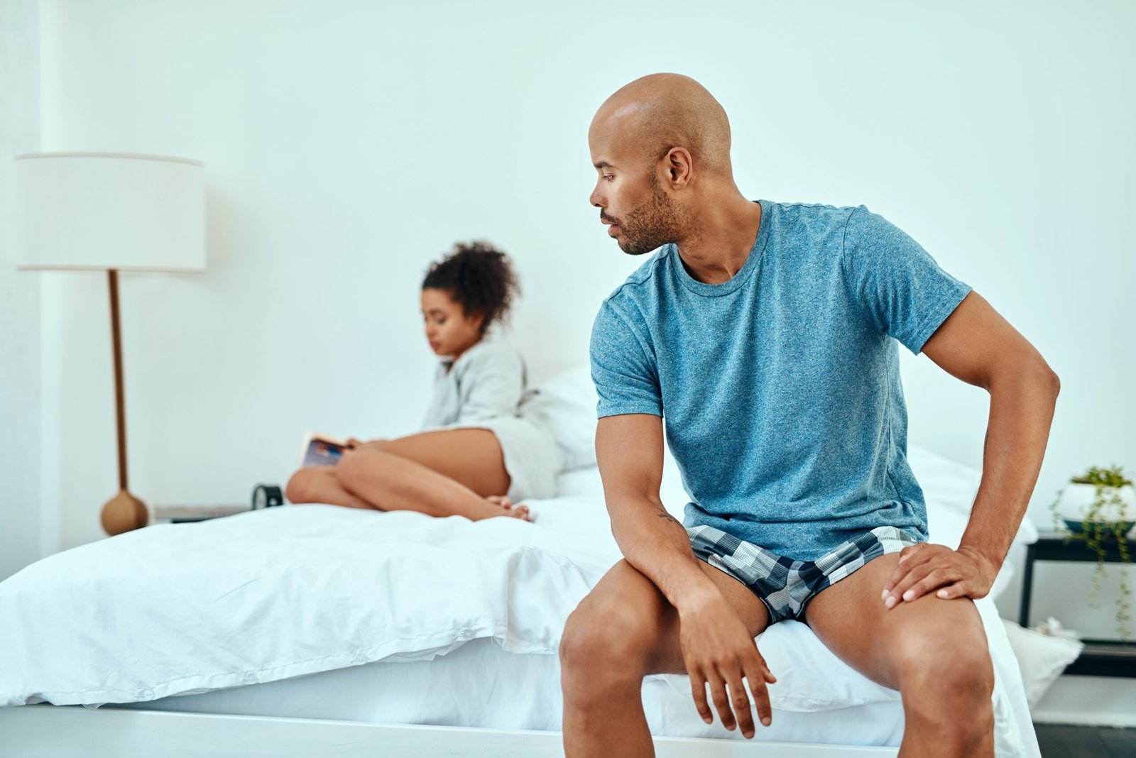 A man sits on the edge of a bed, looking back at a woman who is further behind him, facing away and using her mobile. Both appear thoughtful in a bright, minimalist bedroom, reflecting the quiet impact of erectile dysfunction on their relationship.