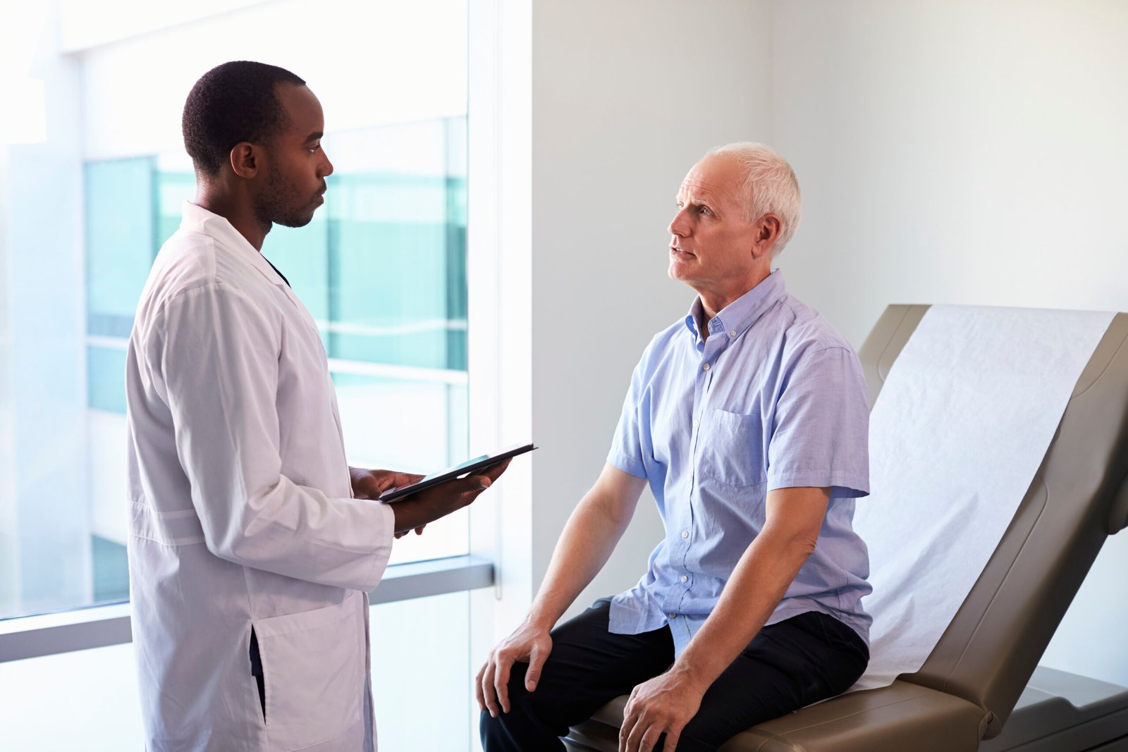 A doctor holding a clipboard talks to an older male patient who is sitting on an examination table in a medical surgery.
