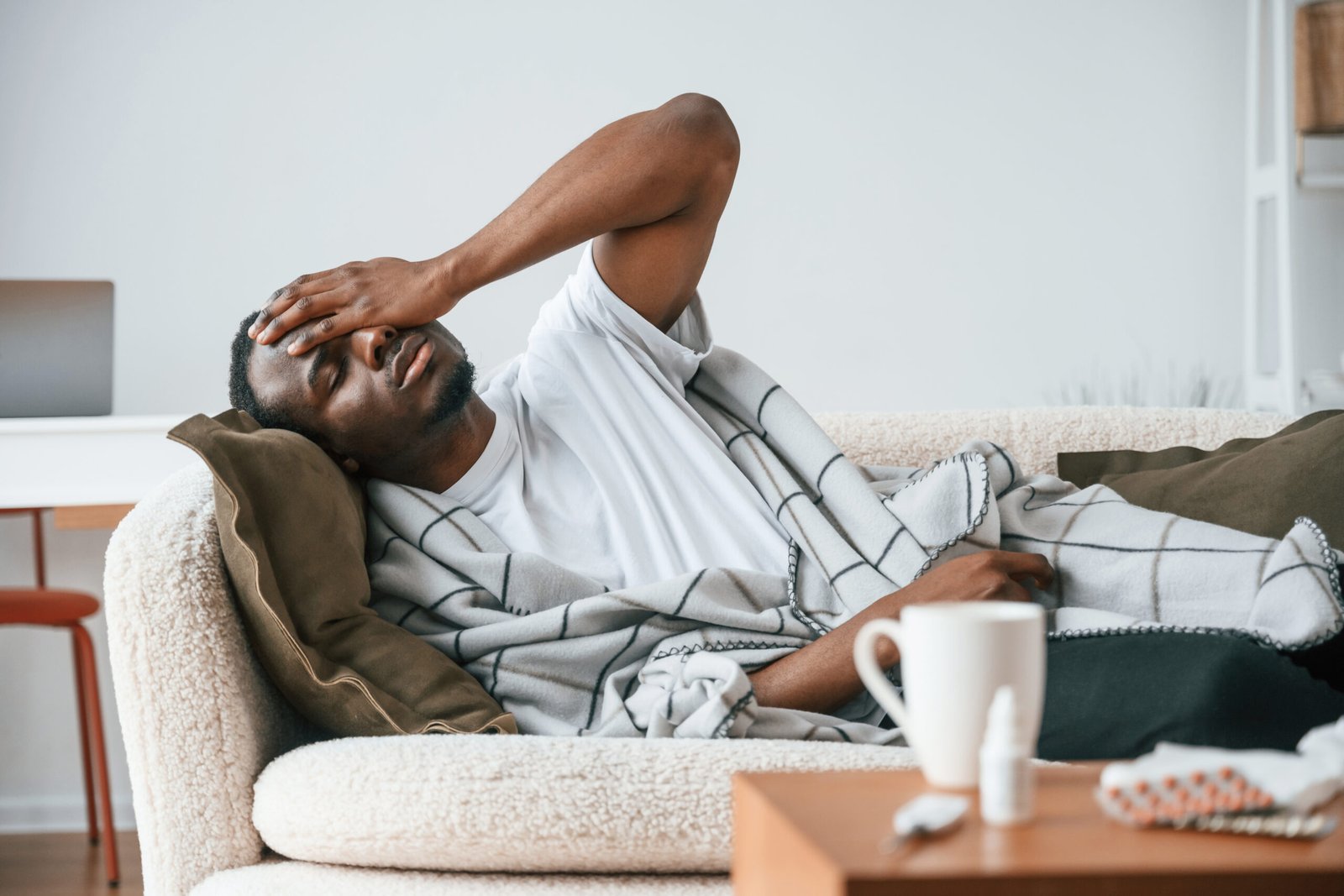 A man lies on a sofa, covered with a blanket, holding his head in discomfort. A mug and a medicine bottle are on the table in front of him, suggesting he may be feeling unwell or has a headache.