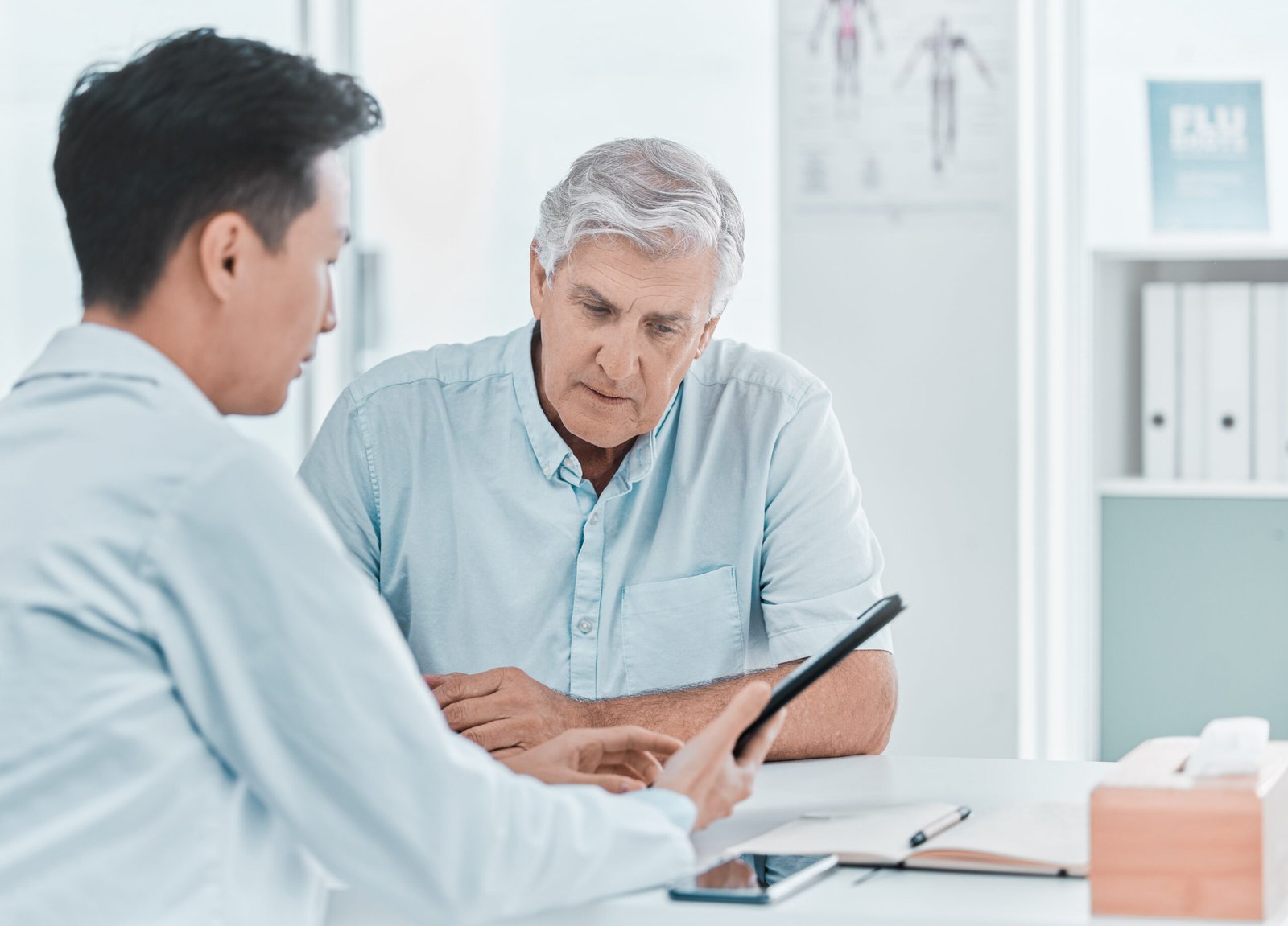 A doctor discusses testosterone and prostate cancer with an older man during a consultation, showing him information on a tablet. Both are seated at a desk, with the patient looking attentively at the screen.