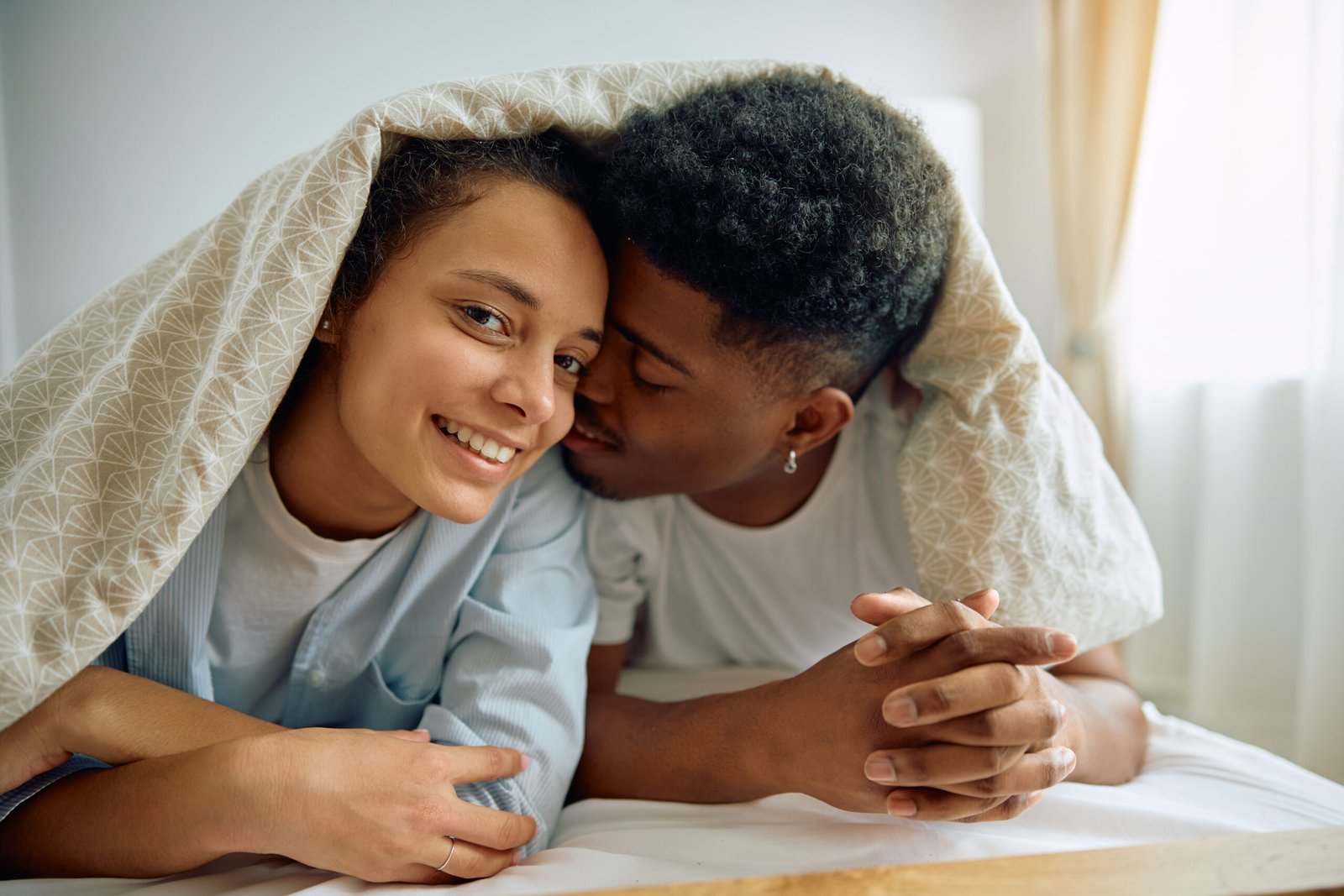 A smiling woman and a man lie close together under a patterned duvet, holding hands on a bed. The woman looks at the camera while the man leans in affectionately, reflecting their normal sex life after prostate surgery in their cosy, sunlit room.