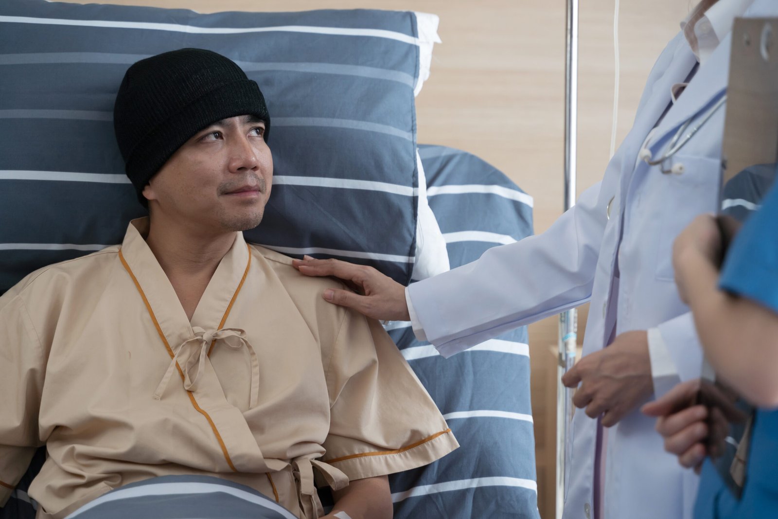 A patient with Metastatic Prostate Cancer wearing a black beanie and hospital gown lies in a hospital bed, looking up at a doctor who gently touches his shoulder whilst another member of medical staff stands nearby.