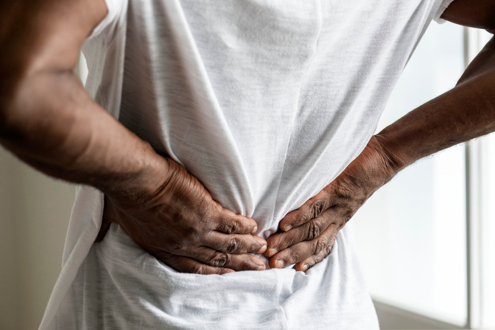 A person wearing a white shirt is standing indoors with their hands pressed against their lower back, appearing to experience backache, which can sometimes be associated with conditions like prostate cancer.