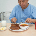 An older man in a hospital gown sits at a table with a large steak, a bowl of soup, a glass of milk, a can of soda, and a small plate of butter cubes in front of him. He is cutting the steak with a knife and fork.