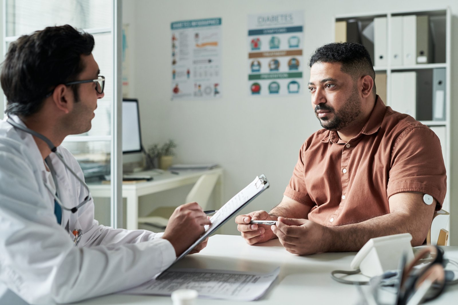 A doctor holding a clipboard talks with a man wearing a glucose monitor on his arm in a medical surgery. The patient listens attentively, with diabetes posters visible on the wall behind them.