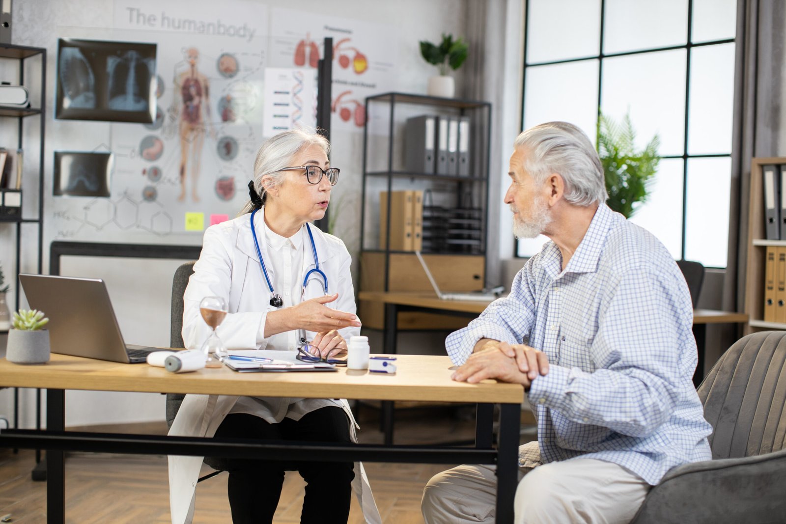 An older female doctor discusses the stages of prostate cancer with an elderly male patient in a medical surgery. They sit opposite each other at a desk with paperwork, a laptop, medicine bottles, and medical charts in the background.