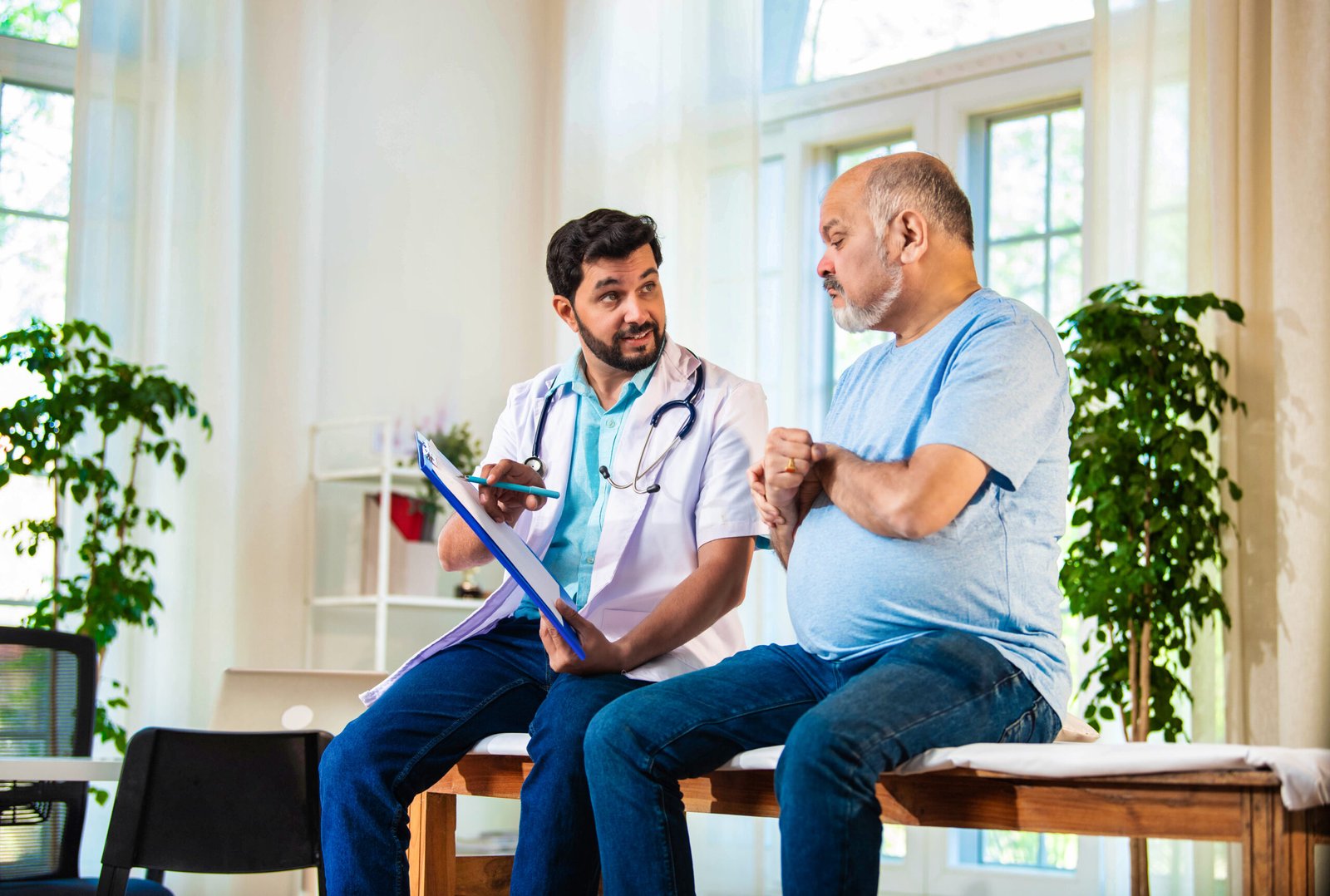 A doctor holding a clipboard discusses metabolic syndrome and prostate cancer with an older man in a light blue shirt, seated on an examination table in a bright, sunlit medical surgery with green plants in the background.
