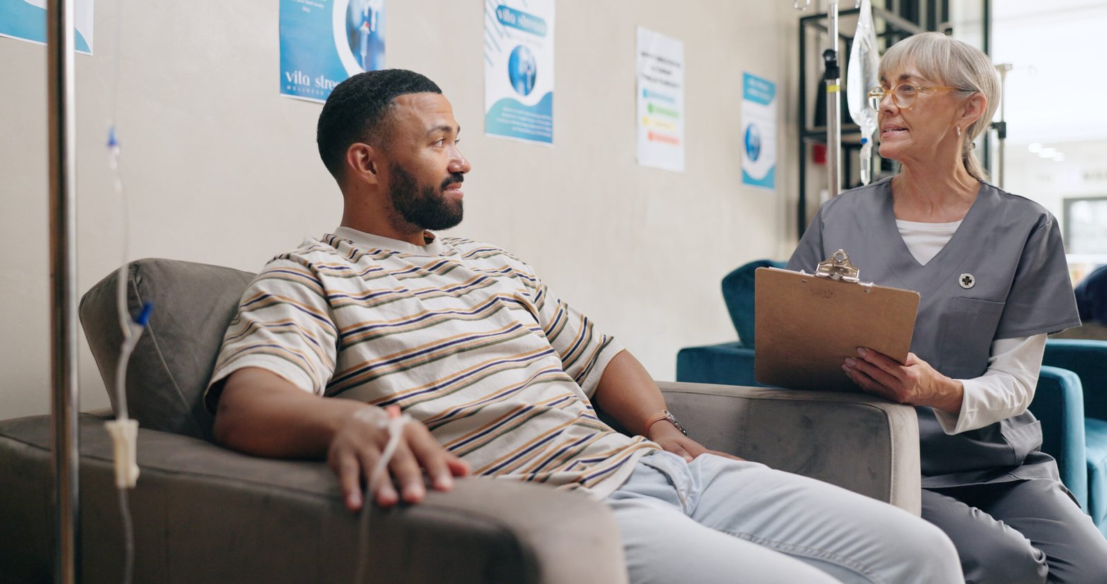 A man sits on a sofa receiving chemotherapy for prostate cancer via drip, while an older woman in scrubs with a clipboard talks to him in a medical setting. Informational posters are visible on the wall behind them.