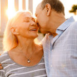 An elderly couple embraces affectionately outdoors at sunset. The woman smiles with closed eyes whilst the man gently kisses her nose. Both appear happy and relaxed, showing love and intimacy despite challenges like Prostate Cancer and Your Sex Life.