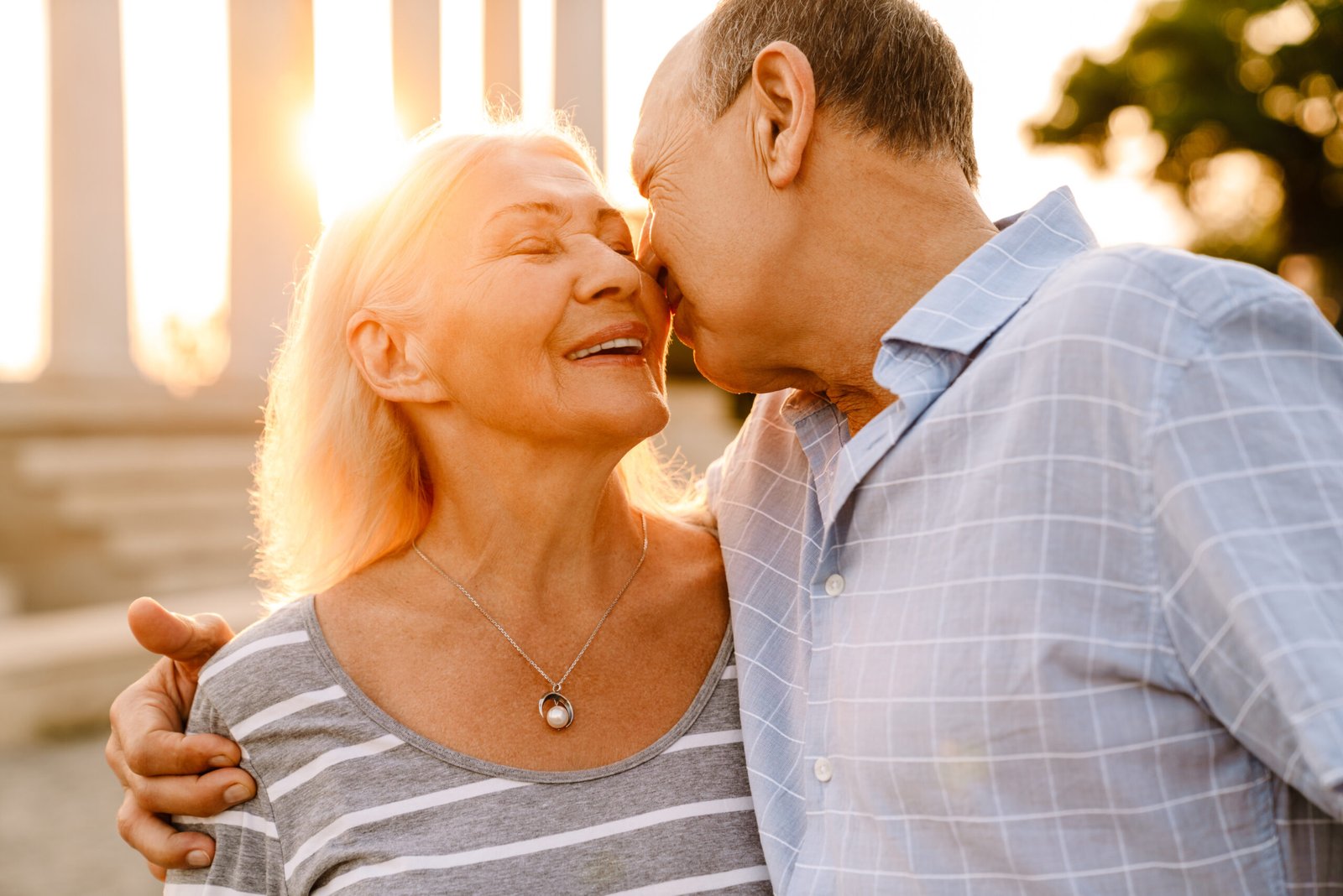An elderly couple embraces affectionately outdoors at sunset. The woman smiles with closed eyes whilst the man gently kisses her nose. Both appear happy and relaxed, showing love and intimacy despite challenges like Prostate Cancer and Your Sex Life.