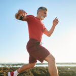 Older man in red sportswear runs energetically outdoors on rocky terrain near the water, listening to music with white earphones under a clear, sunny sky.