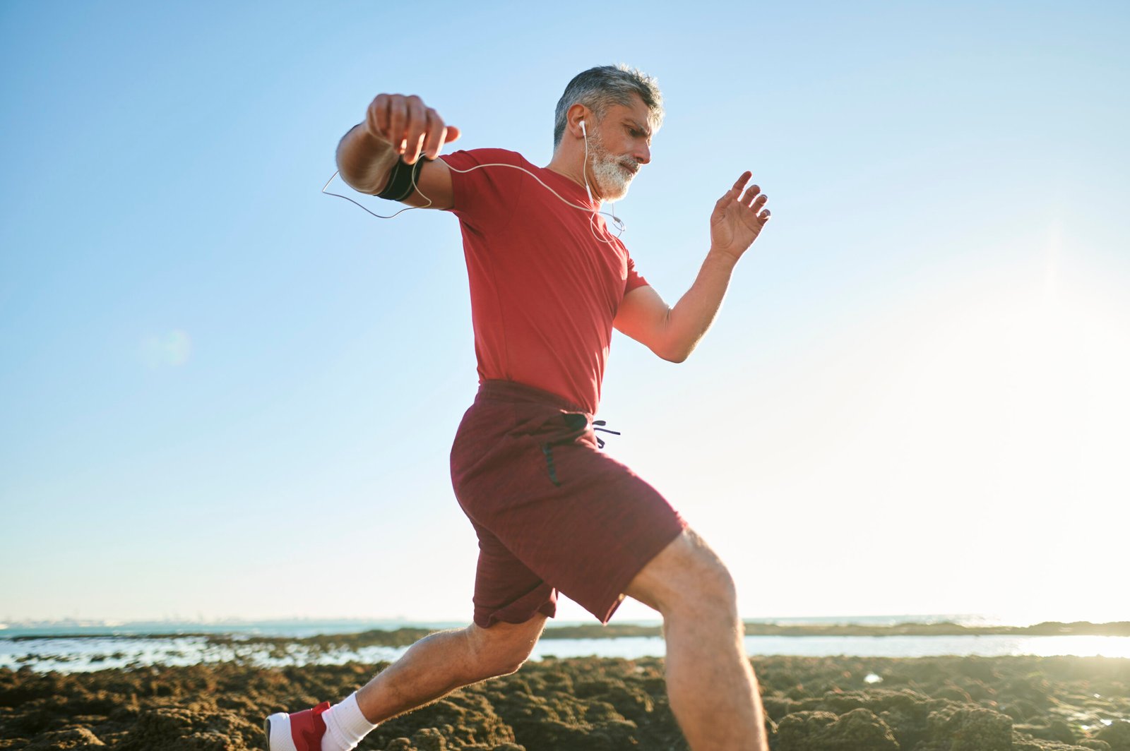 Older man in red sportswear runs energetically outdoors on rocky terrain near the water, listening to music with white earphones under a clear, sunny sky.