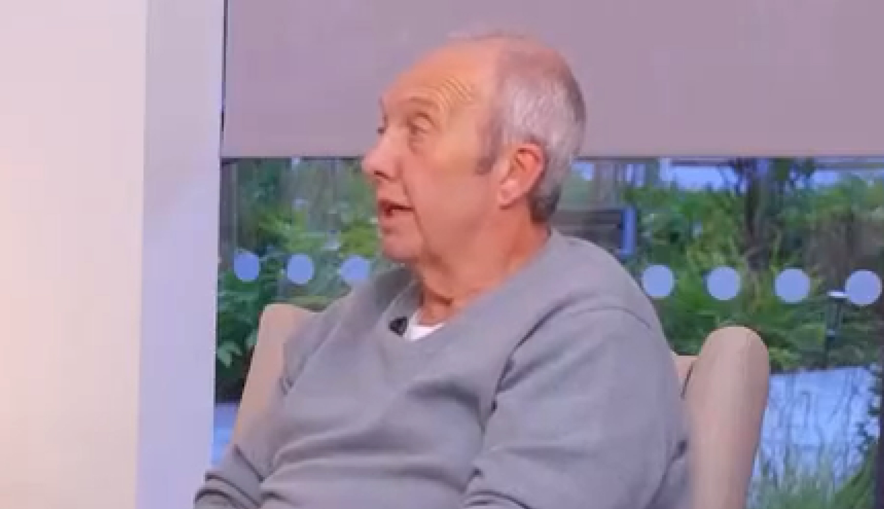 An older man with short grey hair, wearing a light grey jumper, sits indoors on a beige chair, looking to his left. A window and greenery are visible in the background.
