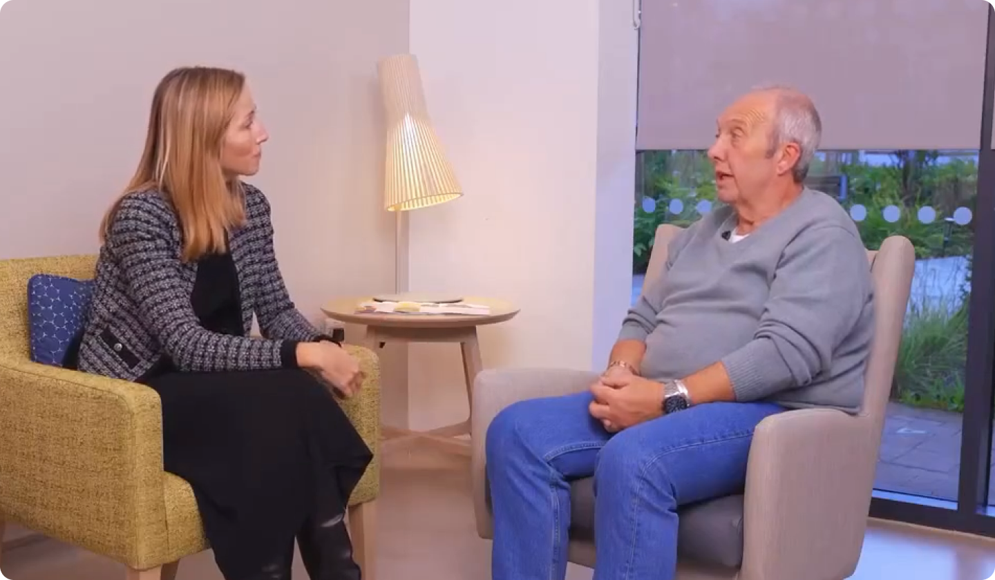 A woman and an older man sit facing each other in armchairs, engaged in conversation in a well-lit room with a lamp and large window in the background.