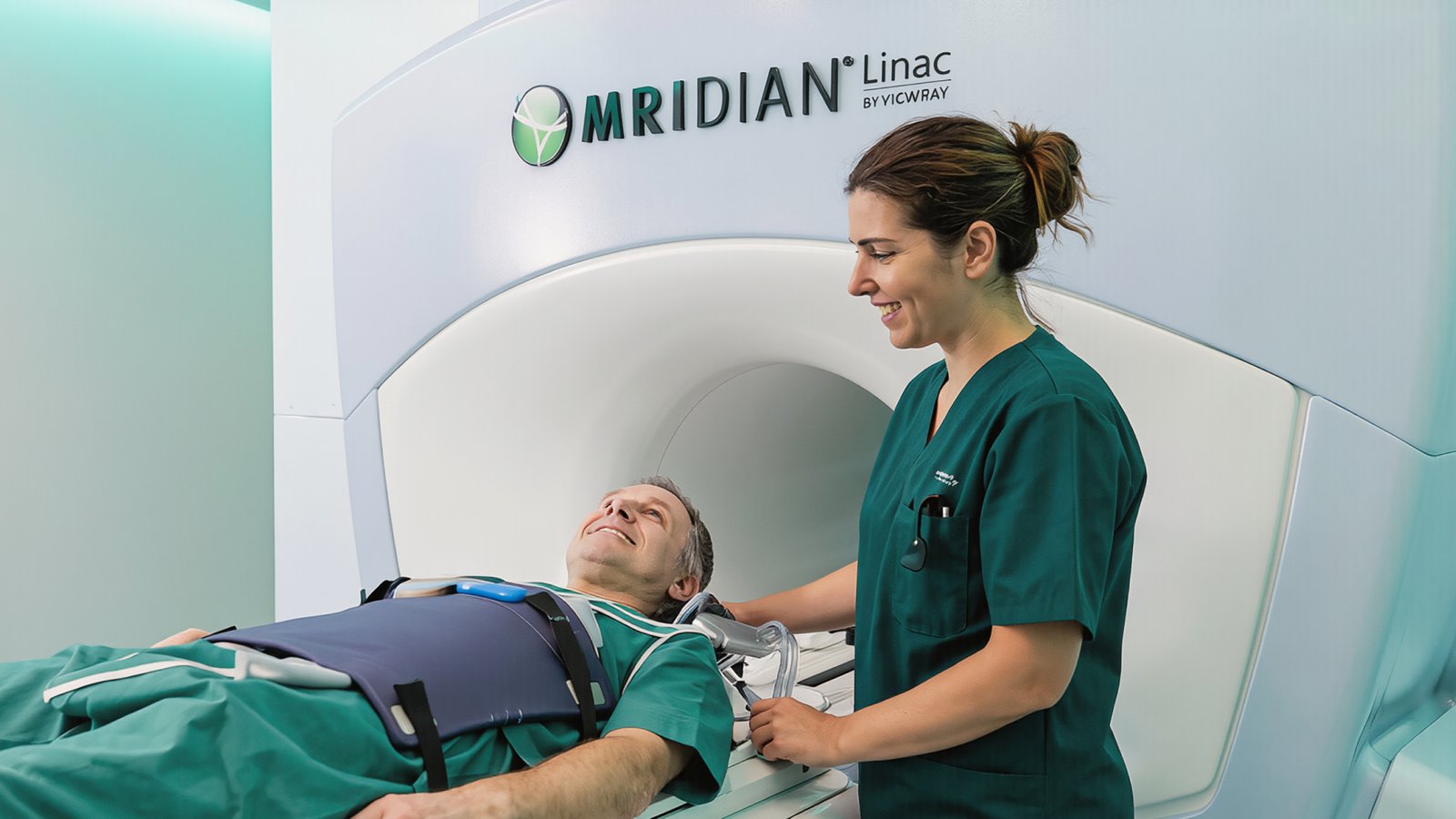 A patient lies on a table inside an MRIdian Linac MRI machine while a healthcare professional in green scrubs stands beside him, smiling and adjusting equipment.