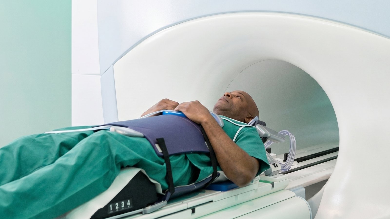 A person in green medical scrubs lies on a table, being moved into an MRI machine for a scan, with medical restraints securing them.