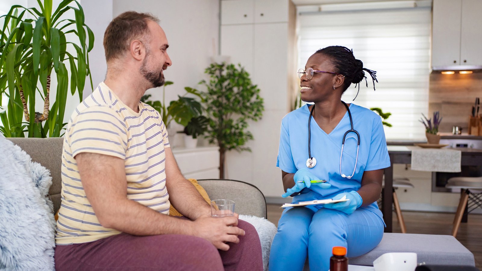 A nurse in blue scrubs smiles whilst talking to a man sitting on a sofa holding a glass of water. They are in a bright living room with plants and medical supplies on the table.
