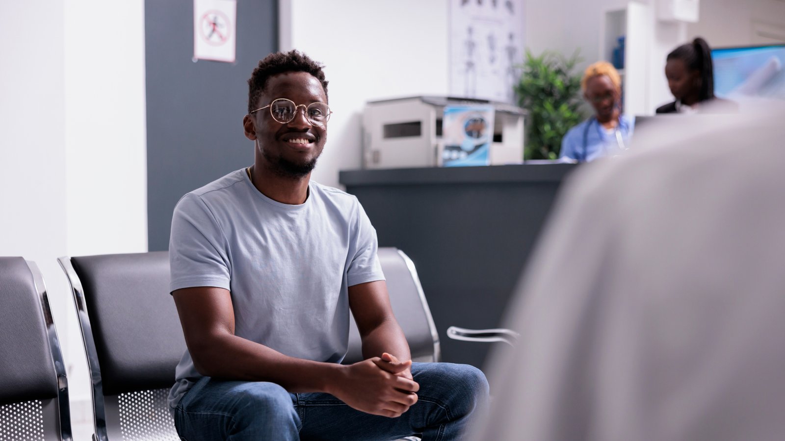A smiling man wearing glasses and a light grey t-shirt sits on a metal chair in a waiting area, with a reception desk and two people in the background.