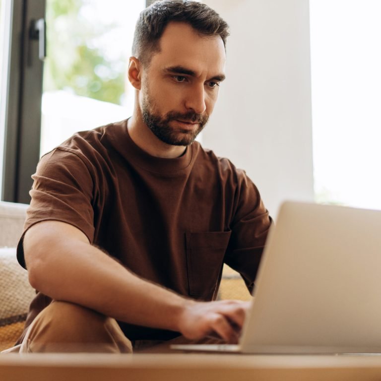 Portrait of attractive smiling Hispanic man wearing stylish eyeglasses and casual clothes sitting on sofa at home using laptop, copywriter typing, ordering. Remote job concept
