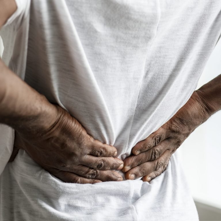 A person wearing a white shirt is standing indoors with their hands pressed against their lower back, appearing to experience backache, which can sometimes be associated with conditions like prostate cancer.