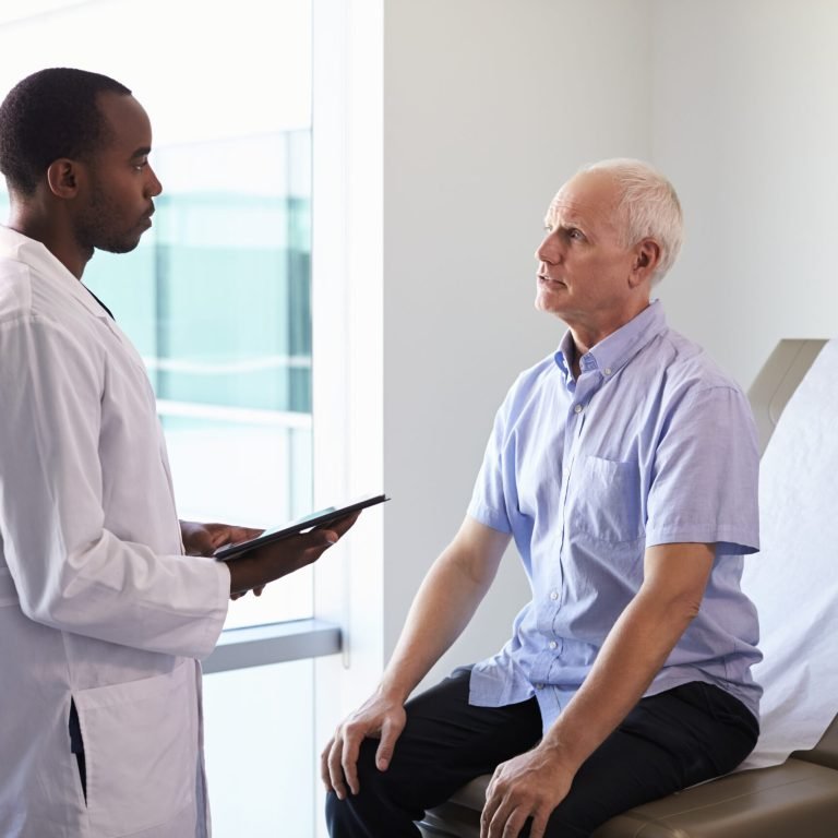 A doctor holding a clipboard talks to an older male patient who is sitting on an examination table in a medical surgery.