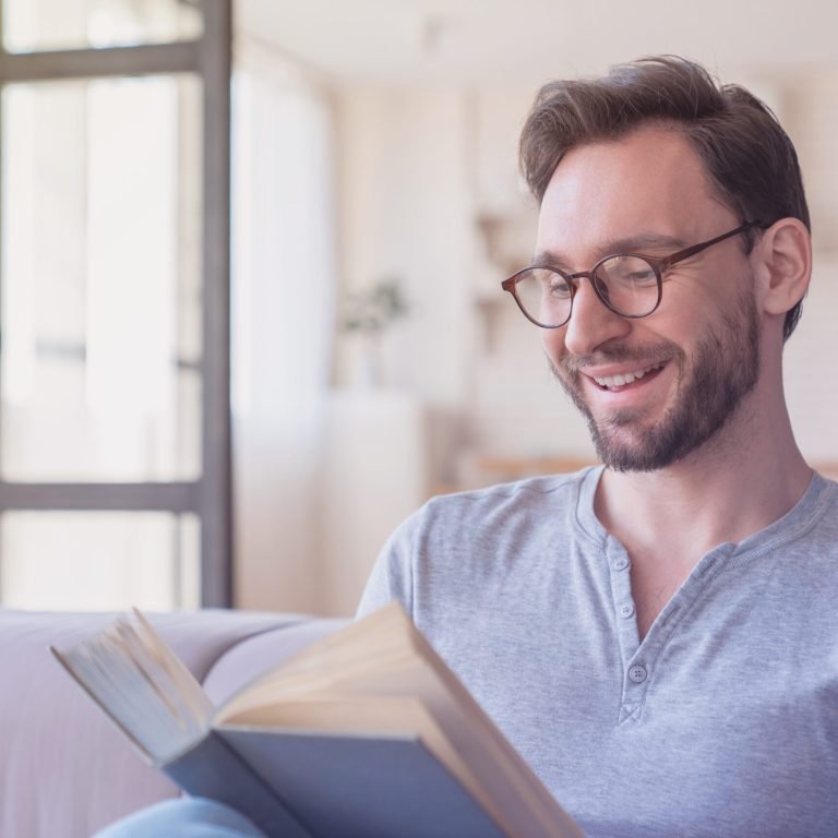 Excited mature caucasian man with eyeglasses reading a book, e-learning, having a break after work at home. Leisure time, weekend and hobby indoors. Adult student resting chilling sitting a couch sofa