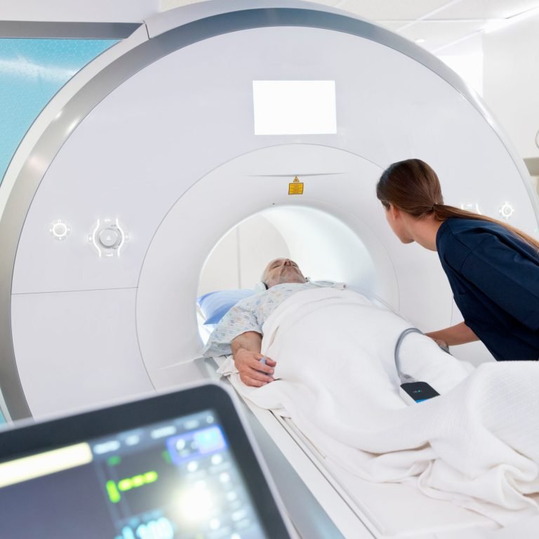 A medical professional assists an elderly patient lying on a bed as they are positioned for an MRI scan to check for prostate cancer, with medical equipment visible in the foreground.