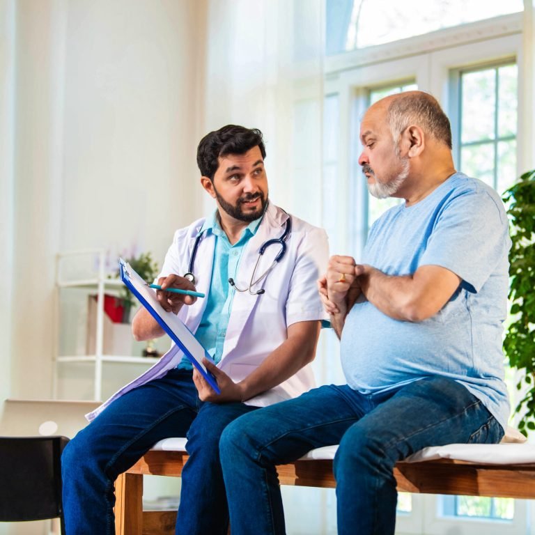 A doctor holding a clipboard discusses metabolic syndrome and prostate cancer with an older man in a light blue shirt, seated on an examination table in a bright, sunlit medical surgery with green plants in the background.