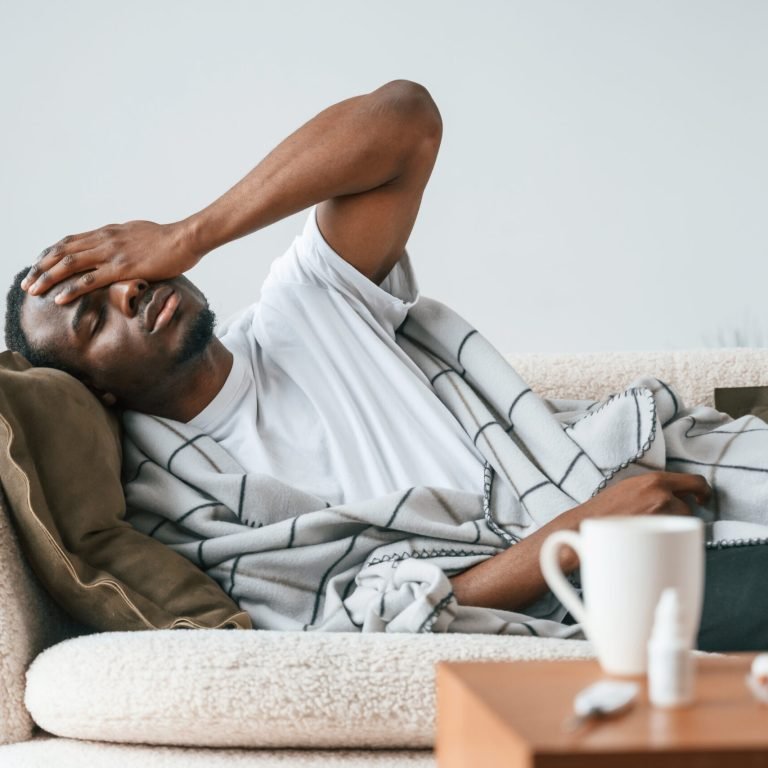 A man lies on a sofa, covered with a blanket, holding his head in discomfort. A mug and a medicine bottle are on the table in front of him, suggesting he may be feeling unwell or has a headache.