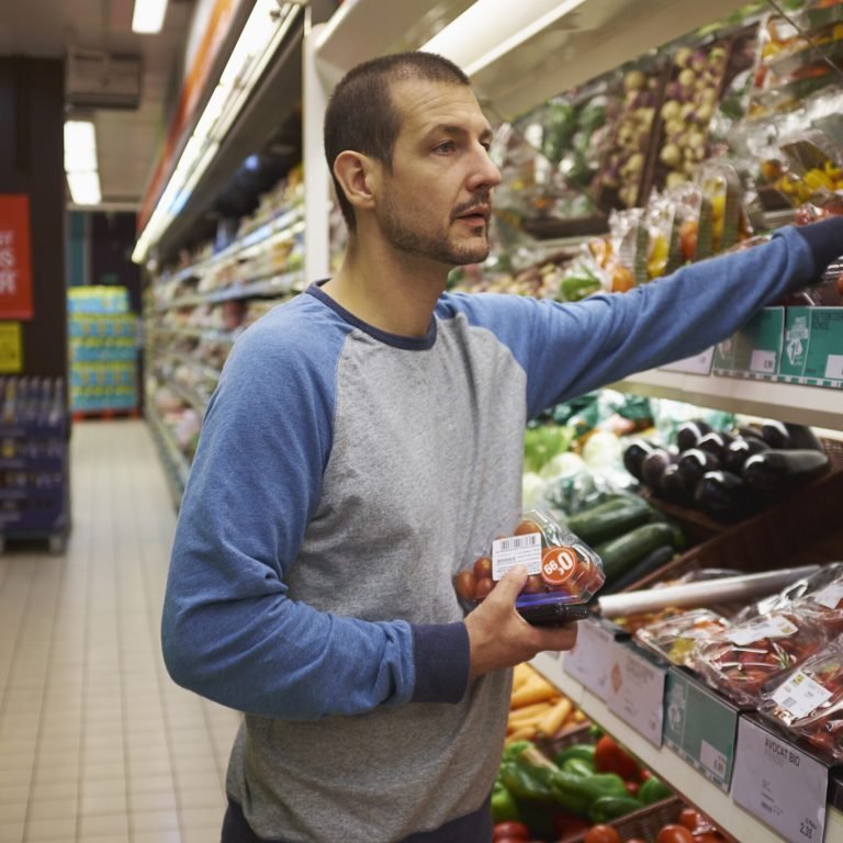 A man wearing a blue and grey long-sleeve shirt shops for fresh vegetables in a supermarket, holding packaged tomatoes while reaching for produce on a shelf.