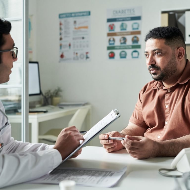 A doctor holding a clipboard talks with a man wearing a glucose monitor on his arm in a medical surgery. The patient listens attentively, with diabetes posters visible on the wall behind them.