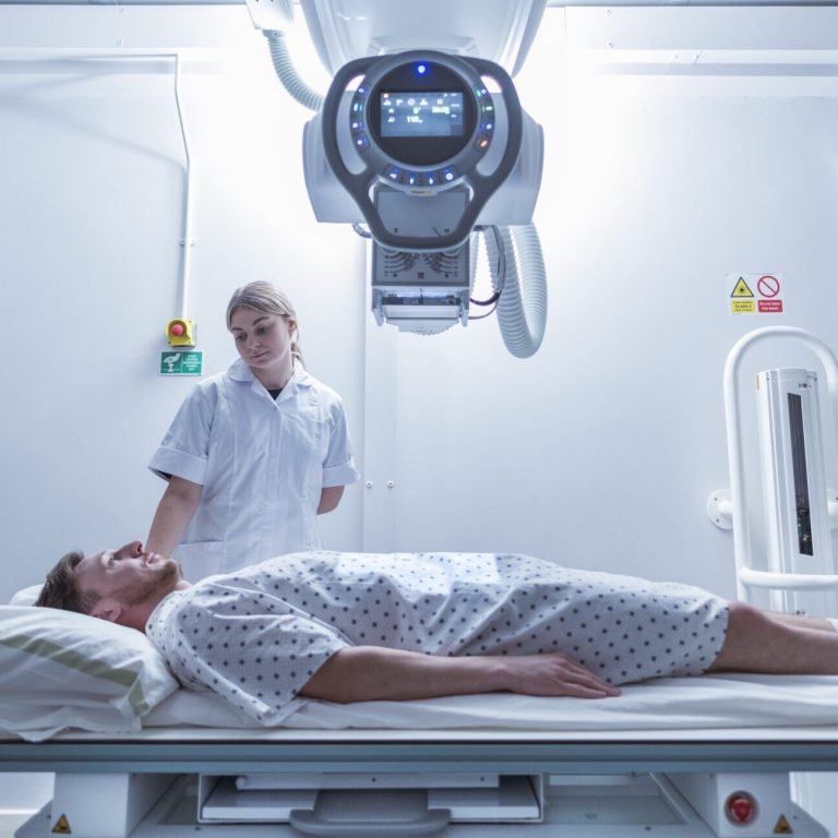 A patient in a hospital gown lies on a bed under a large medical imaging machine, whilst a healthcare worker in a white uniform stands nearby in a bright, modern hospital ward.