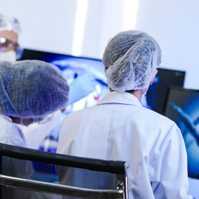 Three scientists wearing lab coats, hairnets, and masks are seated at computer monitors displaying DNA graphics, collaborating on research into treatment options for prostate cancer in a modern laboratory setting.