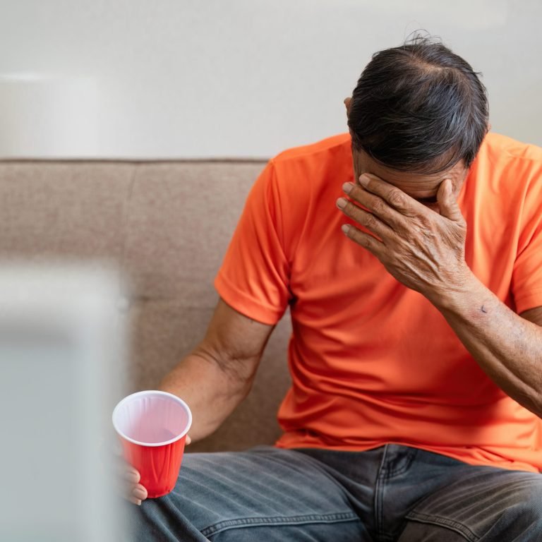 A man in an orange shirt sits on a sofa, holding a red cup in one hand and covering his face with the other, appearing distressed—possibly after receiving news about prostate cancer. Another person stands nearby, partially visible.