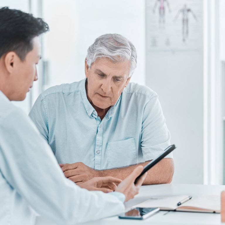 A doctor discusses testosterone and prostate cancer with an older man during a consultation, showing him information on a tablet. Both are seated at a desk, with the patient looking attentively at the screen.