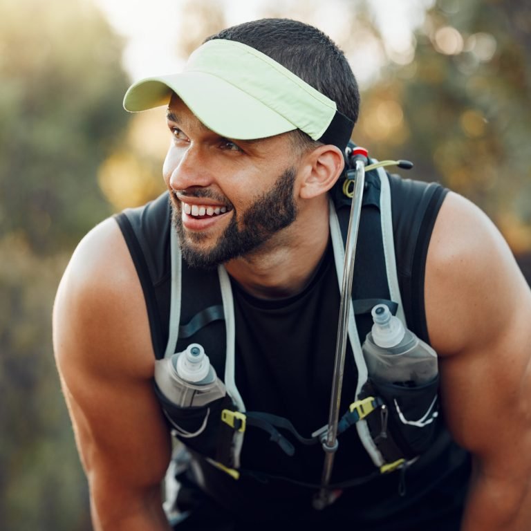 A smiling man with a beard wears a sun visor and hydration vest with water bottles, leaning forward outdoors on a sunny day after running or hiking—staying active while exploring treatment options for prostate cancer. Trees and sunlight fill the background.