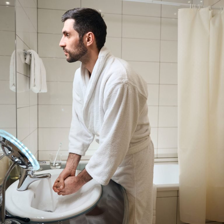 A man in a white dressing gown stands at a bathroom sink, washing his hands. The bathroom has white tiled walls, a mirror, a towel, and a shower curtain in the background.