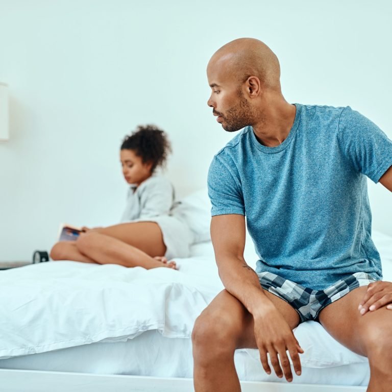 A man sits on the edge of a bed, looking back at a woman who is further behind him, facing away and using her mobile. Both appear thoughtful in a bright, minimalist bedroom, reflecting the quiet impact of erectile dysfunction on their relationship.