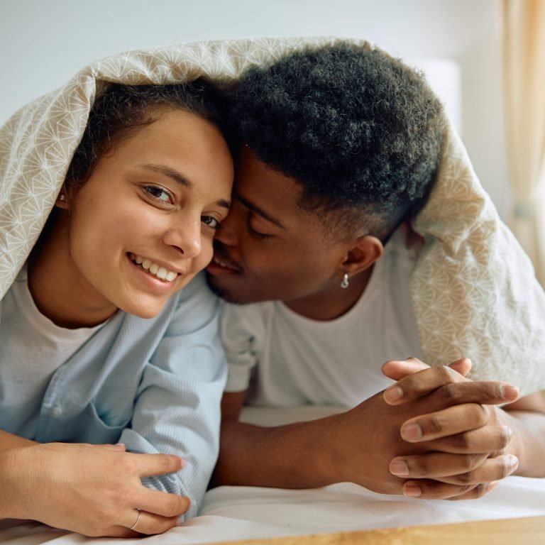 A smiling woman and a man lie close together under a patterned duvet, holding hands on a bed. The woman looks at the camera while the man leans in affectionately, reflecting their normal sex life after prostate surgery in their cosy, sunlit room.